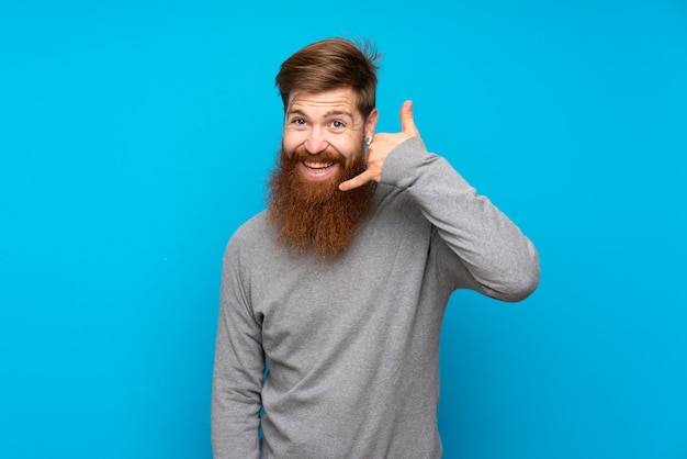 Homme rousse à longue barbe sur bleu isolé faisant geste de téléphone. Rappelez-moi signe