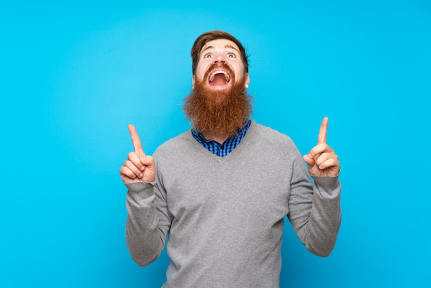 Homme rousse avec longue barbe sur bleu isolé surpris et pointant vers le haut