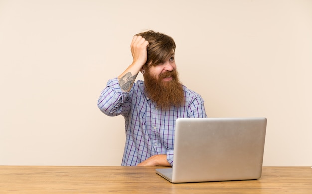 Homme rousse avec une longue barbe dans une table avec un ordinateur portable ayant des doutes et avec une expression du visage confuse