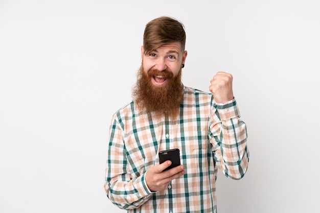 Homme rousse avec longue barbe sur mur blanc isolé avec téléphone en position de victoire