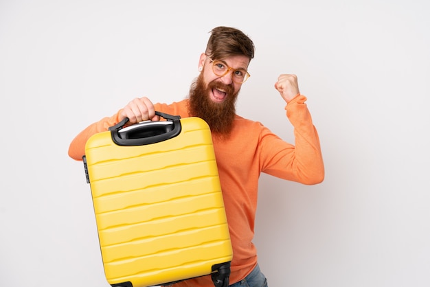 Homme rousse avec longue barbe sur mur blanc isolé en vacances avec valise de voyage et un chapeau