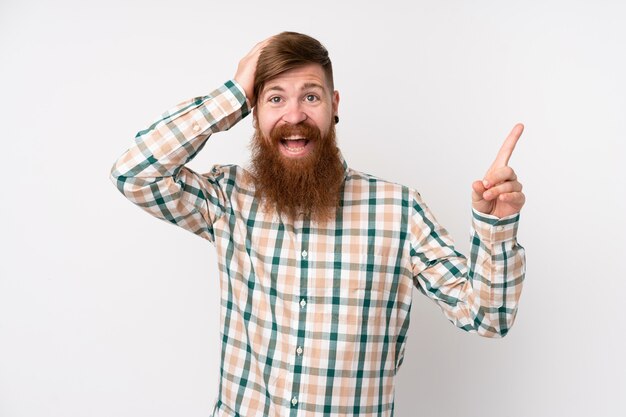 Homme rousse avec longue barbe sur mur blanc surpris et pointant le doigt sur le côté