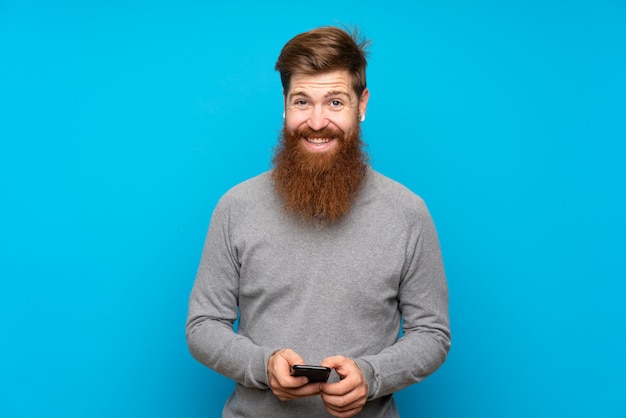 Homme rousse à longue barbe sur mur bleu en envoyant un message avec le mobile