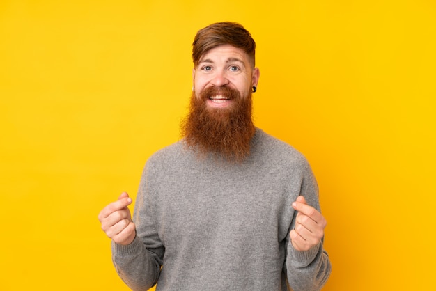 Homme rousse avec une longue barbe sur un mur jaune isolé faisant un geste d'argent