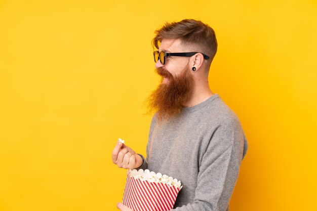 Homme rousse avec une longue barbe sur un mur jaune isolé avec des lunettes 3d et tenant un grand seau de pop-corn