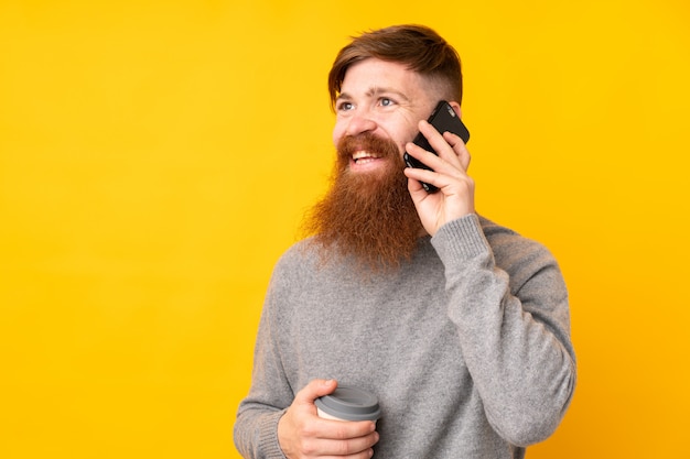 Homme rousse avec une longue barbe sur un mur jaune isolé tenant du café à emporter et un mobile