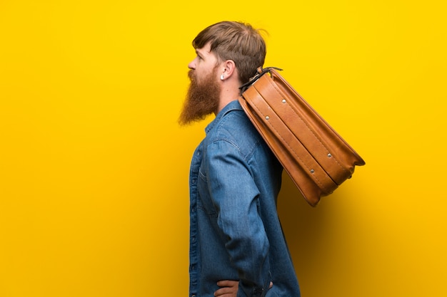 Homme rousse avec une longue barbe sur un mur jaune isolé, tenant une mallette vintage