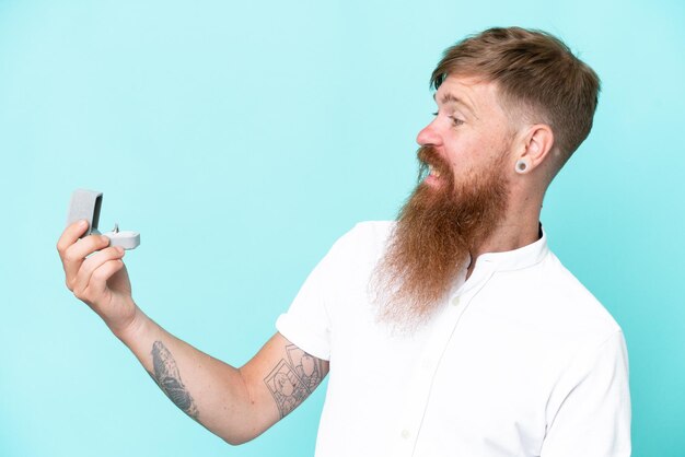 Homme rousse avec une longue barbe tenant une bague de fiançailles isolée sur fond bleu avec une expression heureuse