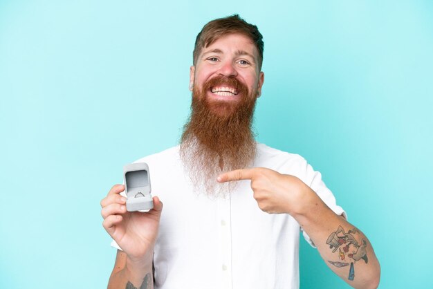 Homme rousse avec une longue barbe tenant une bague de fiançailles isolée sur fond bleu et la pointant