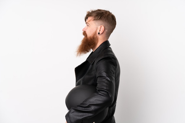 Homme rousse à longue barbe tenant un casque de moto sur un mur blanc isolé à la recherche sur le côté