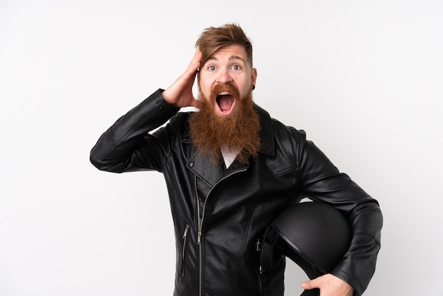 Homme rousse à longue barbe tenant un casque de moto sur un mur blanc isolé surpris et pointant le doigt sur le côté