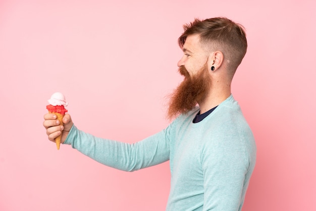 Homme rousse à longue barbe tenant une glace au cornet sur un mur rose isolé avec une expression heureuse