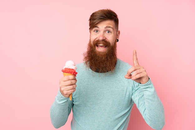 Homme rousse à longue barbe tenant une glace au cornet sur un mur rose isolé pointant vers le haut une excellente idée