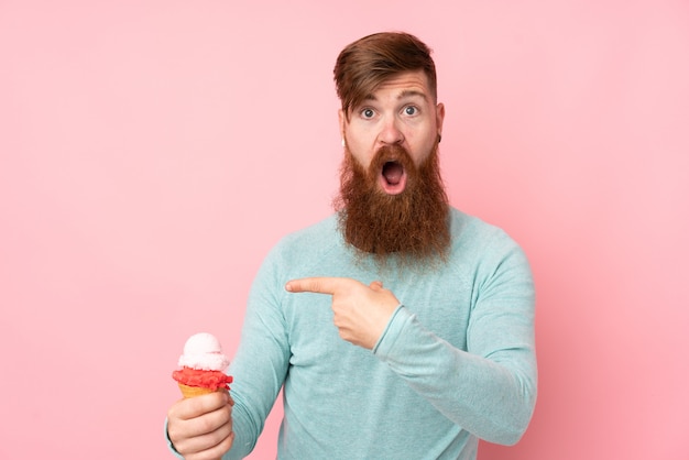 Homme rousse à longue barbe tenant une glace au cornet sur un mur rose surpris et pointant le doigt sur le côté