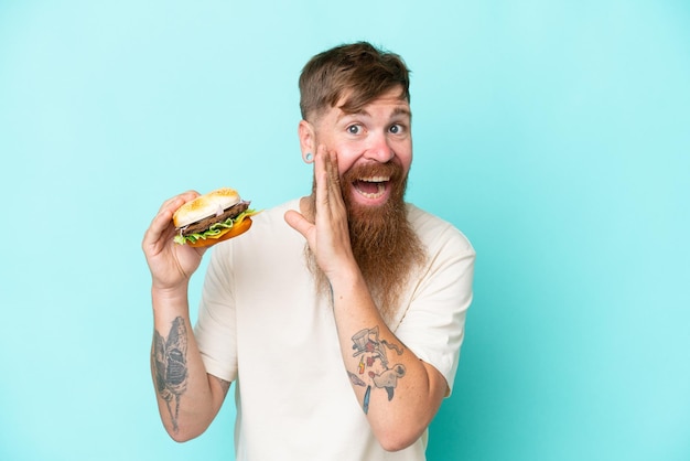 Homme rousse avec une longue barbe tenant un hamburger isolé sur fond bleu chuchotant quelque chose