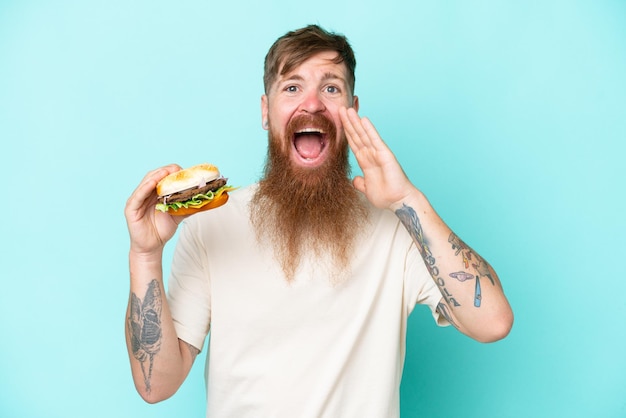 Homme rousse avec une longue barbe tenant un hamburger isolé sur fond bleu criant avec la bouche grande ouverte