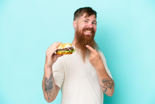 Homme rousse avec une longue barbe tenant un hamburger isolé sur fond bleu et le pointant