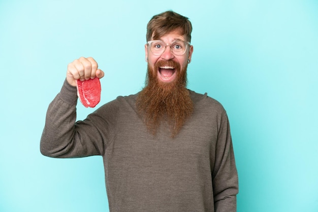 Homme rousse avec une longue barbe tenant un morceau de viande isolé sur fond bleu avec une expression faciale surprise et choquée