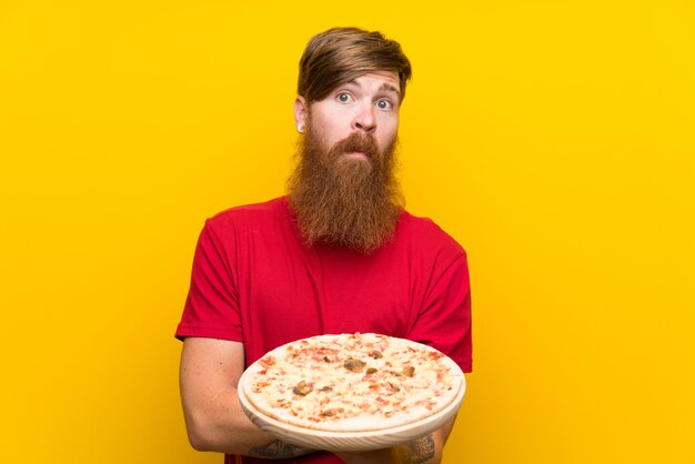 Homme rousse avec une longue barbe tenant une pizza sur un mur jaune isolé, faisant un geste de doute tout en soulevant les épaules