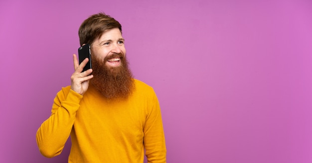 Homme rousse avec une longue barbe sur violet isolé gardant une conversation avec le téléphone mobile