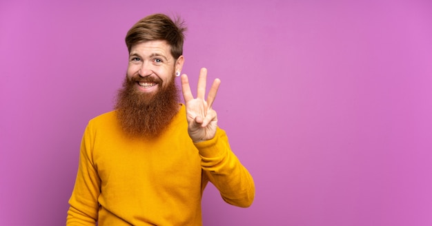 Homme rousse avec une longue barbe sur violet isolé heureux et en comptant trois avec les doigts