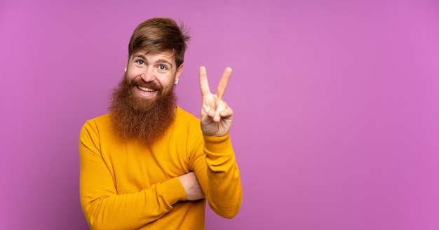 Homme rousse avec longue barbe sur violet isolé souriant et montrant le signe de la victoire