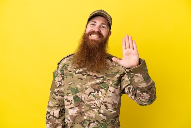 Homme rousse militaire sur isolé sur fond jaune saluant avec la main avec une expression heureuse