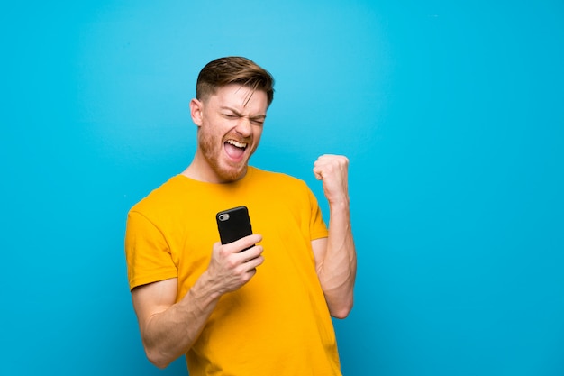 Homme rousse sur un mur bleu avec téléphone en position de victoire