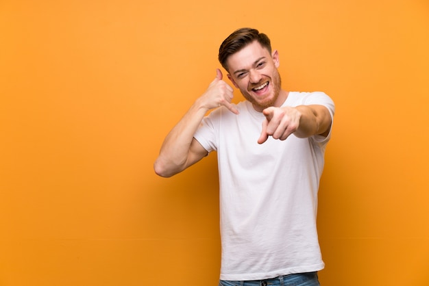 Homme Rousse Sur Un Mur Brun Faisant Un Geste De Téléphone Et Pointant Le Devant