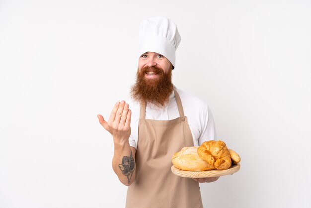 Homme rousse en uniforme de chef. Boulanger mâle tenant une table avec plusieurs pains invitant à venir avec la main.