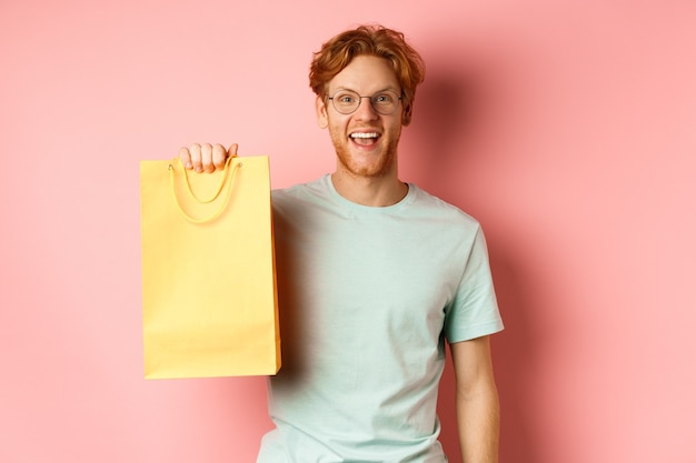 Homme roux gai en t-shirt, montrant un sac à provisions et souriant, debout sur fond rose.