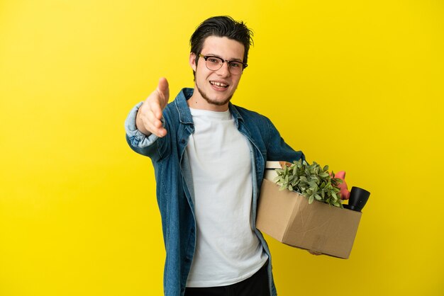 Homme russe faisant un mouvement en ramassant une boîte pleine de choses isolées sur fond jaune se serrant la main pour conclure une bonne affaire