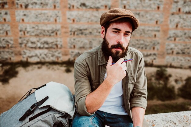 Un homme avec un sac à dos tenant un crayon et un livre alors qu'il est assis à l'extérieur.