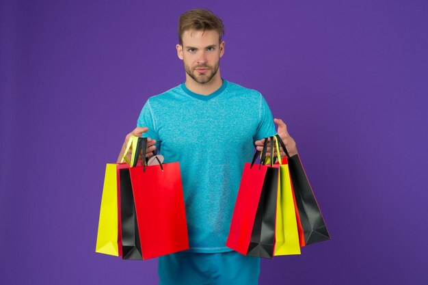 Homme avec des sacs à provisions sur fond violet. Macho avec des sacs en papier colorés. Acheteur de mode en t-shirt bleu décontracté. Préparation et célébration des vacances. Shopping ou vente et cyber lundi.