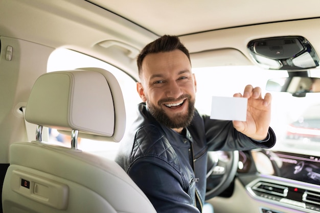 Un homme satisfait avec un sourire sur son visage est assis dans le siège du conducteur et montre une carte de visite au