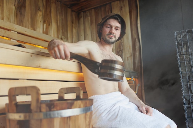 L'homme se détend dans un sauna finlandais et tire de l'eau d'un seau en bois avec une pelle