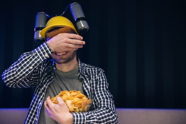 L'homme se repose à la maison et regarde des émissions de télévision ou des nouvelles sportives sur l'écran de télévision avec un casque de bière sur la tête mange des chips