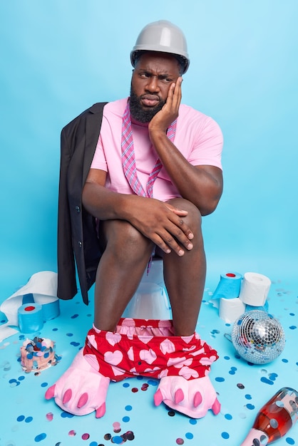 l'homme se sent fatigué après la fête pose sur la cuvette des toilettes souffre de diarrhée porte des pantoufles de t-shirt de casque pose dans des toilettes en désordre après la célébration