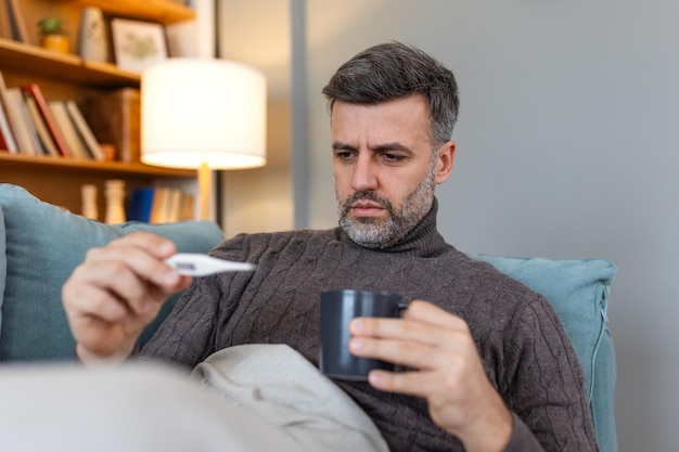 Photo un homme se sent malade allongé dans le lit et regardant le thermomètre un homme malade étendu sur le canapé vérifiant sa température sous une couverture à la maison dans le salon