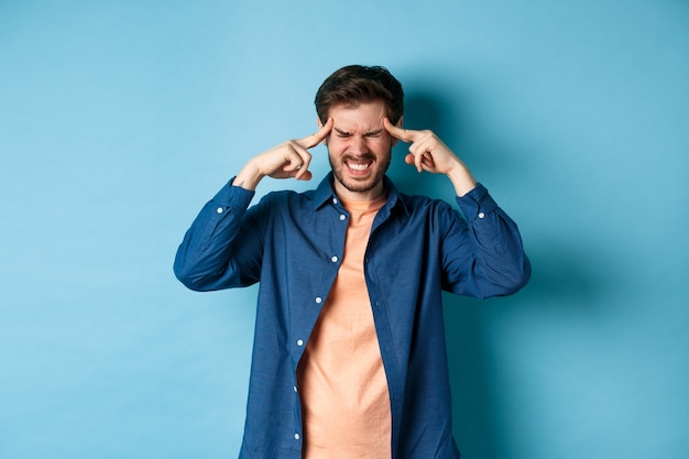 Homme se sentant malade, touchant la tête et grimaçant de migraine douloureuse, ayant mal à la tête ou se sentant étourdi, debout sur fond bleu.