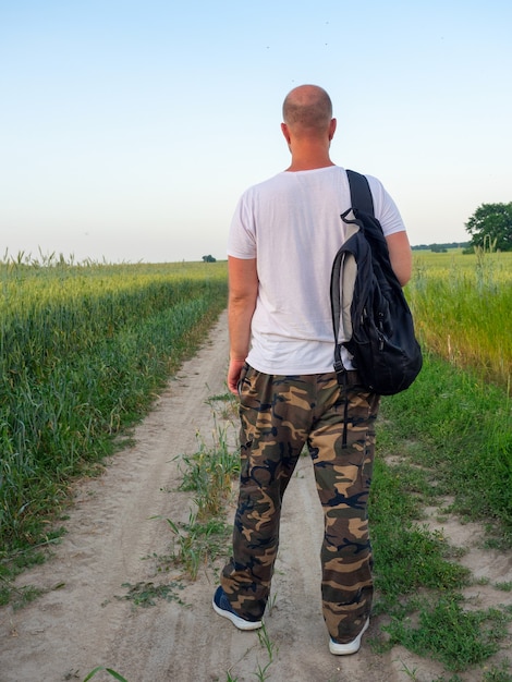 Un homme se tient sur un chemin dans un champ avec un sac à dos noir dos à la caméra. Paysage du soir, concept de voyage