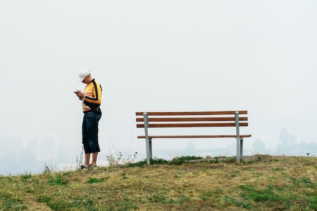 Un homme se tient sur une colline avec un téléphone dans ses mains sur un fond de brouillard épais Un banc vide et un jeune homme debout à côté Le concept de solitude et de nostalgie