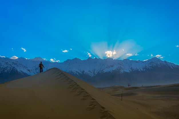 Un homme se tient sur une dune de sable dans le désert avec des montagnes en arrière-plan.