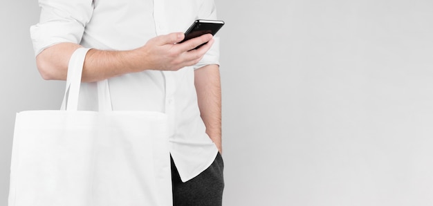 Un homme se tient sur un fond blanc, lit les nouvelles au téléphone et tient un sac écologique en lin sur son coude. Concept d'écologie