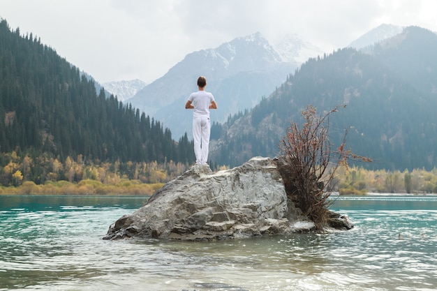 Un homme se tient sur une pierre au centre d'un lac de montagne et pratique le yoga. Pose Vrikshasana.