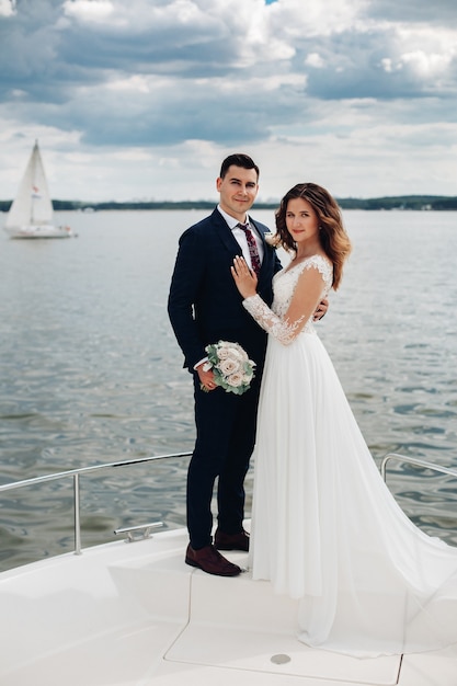 Homme séduisant en costume noir et chemise blanche avec une belle femme en longue robe blanche pose pour la caméra sur un yacht