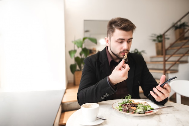 Homme séduisant dans un costume est assis dans un restaurant confortable, manger de la salade avec une assiette et à l'aide d'un smartphone