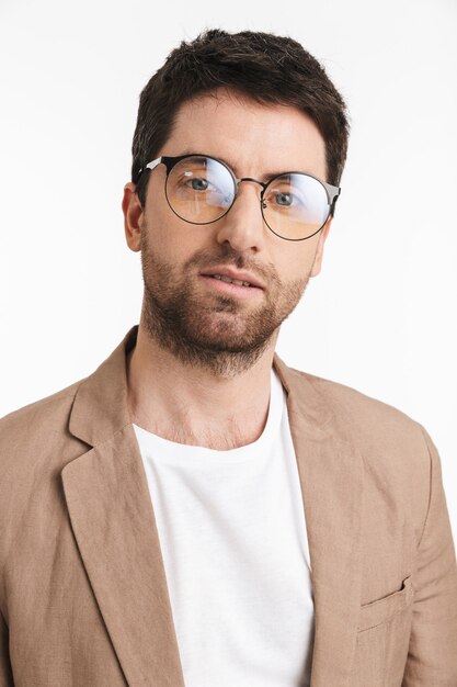 homme séduisant avec du chaume portant une veste et des lunettes posant à l'avant isolé sur un mur blanc