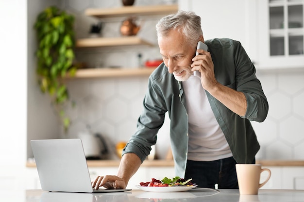 Homme senior d'affaires à distance utilisant un téléphone portable et travaillant sur un ordinateur portable dans la cuisine