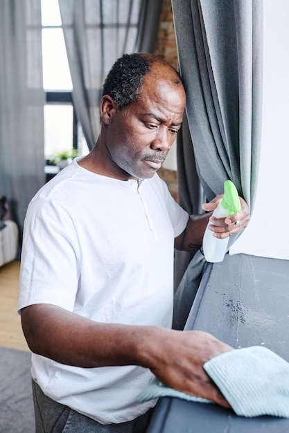 Homme senior afro-américain sérieux en t-shirt blanc saupoudrant le rebord de la fenêtre gris